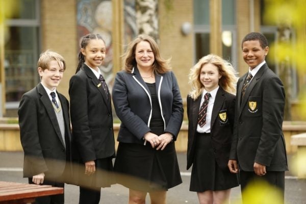 An adult woman stands with four school children wearing black blazers and uniforms outside a school building