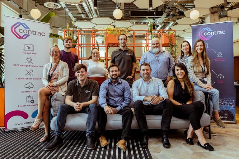 Team photo of diverse professionals posing in front of Contrac branded banners in a modern office setting.