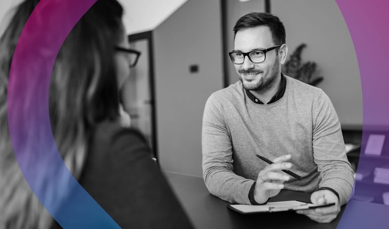 Man wearing glasses and gray sweater smiles while holding a tablet in an office setting with purple graphic design elements
