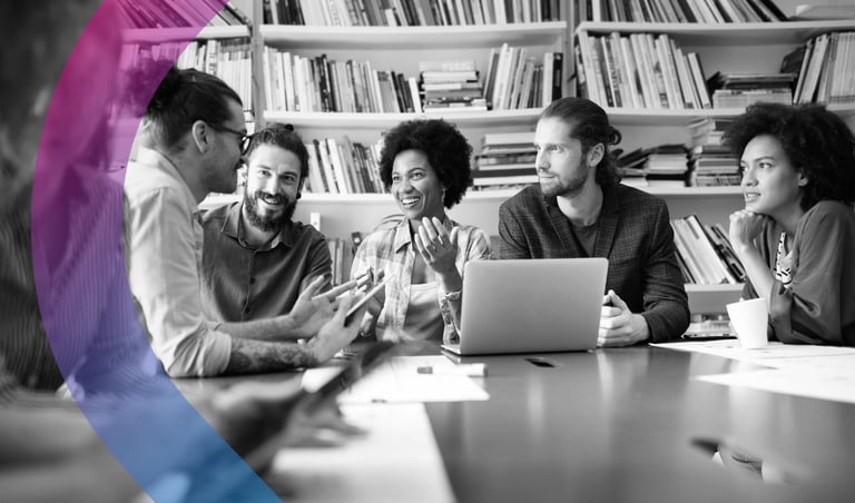 Five diverse professionals in discussion around a table with a laptop and bookshelves in background during a team meeting.